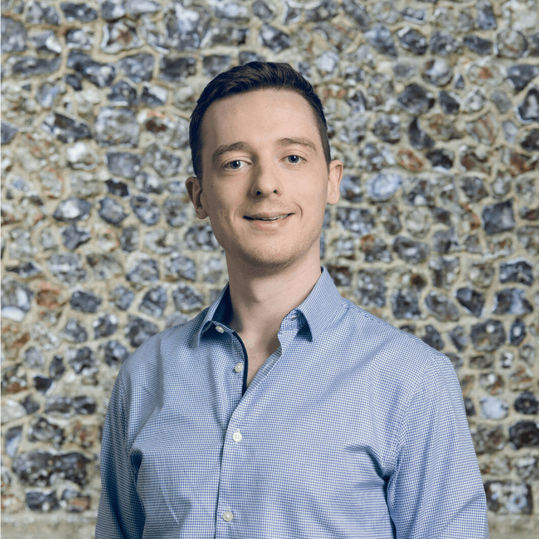A person in a light blue button-up shirt stands against a textured stone wall background, smiling slightly at the camera.