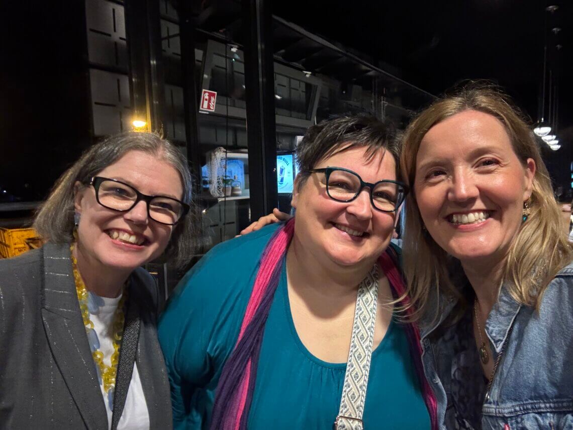 Three smiling women taking a selfie in an indoor setting, with a glass wall in the background and dim lighting.