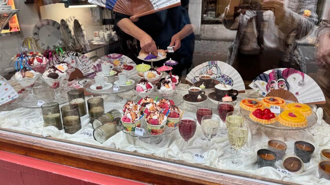 A shop window displaying an array of colorful desserts, including ice cream sundaes, cakes, and drinks. Elegant ceramic plates and decorative fans are also showcased, with prices visible on some items.