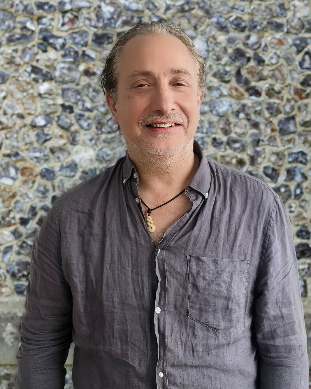 Individual in a grey shirt stands smiling against a textured stone wall.