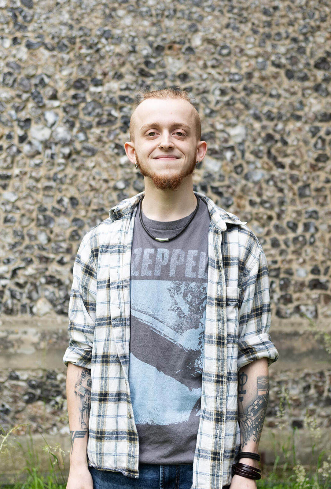 Person smiling in a plaid shirt and band t-shirt, standing against a stone wall.