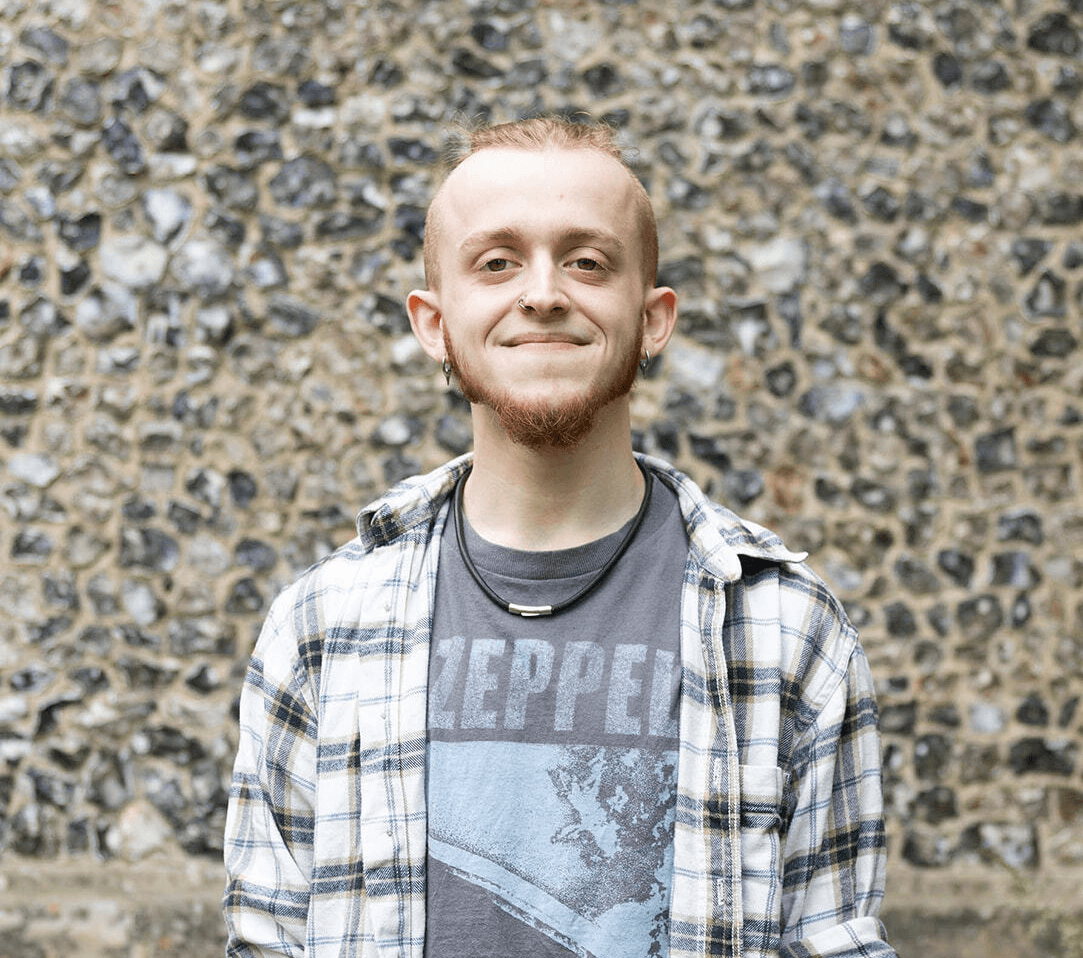 A person with a relaxed smile stands against a textured stone wall, wearing casual clothing.